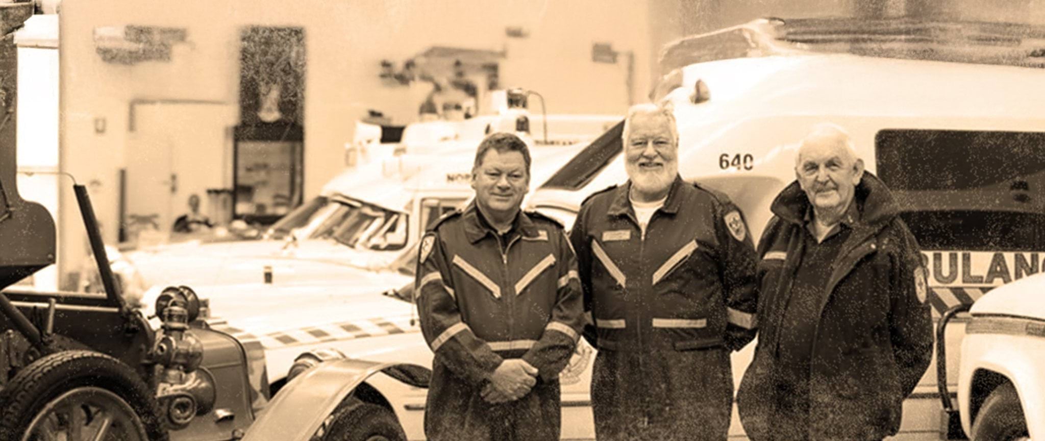 Three retired paramedics and first responders standing in front of several vintage ambulance vehicles parked inside a garage.