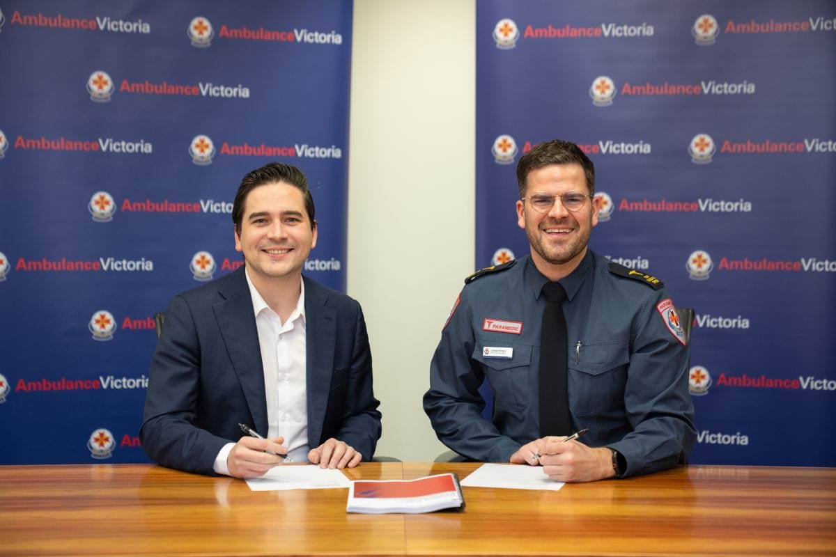 Two smiling men pose at a desk, one in a suit and the other in a parmedic formal uniform
