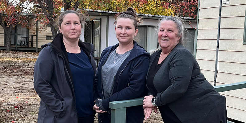 Three female paramedics smiling and standing outside the porch of a cabin with trees in autumn colours in the background.