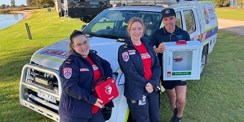 Two female paramedics and a man, who is the owner of Lake Boga Caravan Park, are standing beside an Ambulance Victoria vehicle parked beside Lake Boga. The female paramedic is holding the new automated external defibrillator device and the man is holding the outer display casing for the device.