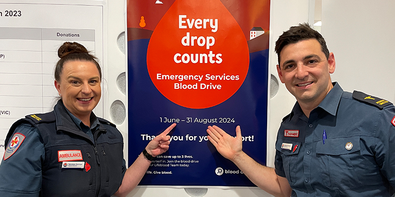 Two Ambulance Victoria staff pointing to an Emergency Services Blood Drive campaign poster.