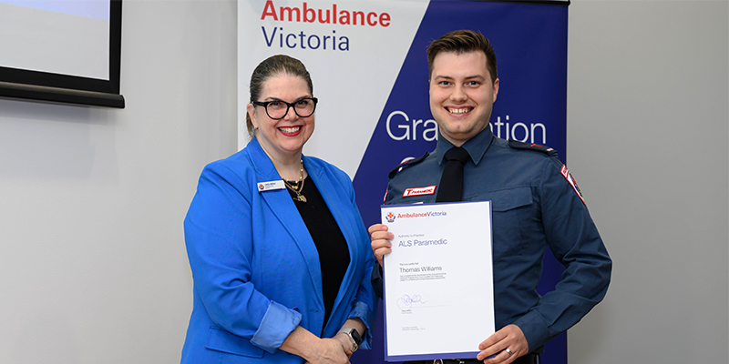 A male graduate paramedic is holding his graduation certificate. He is standing beside Ambulance Victoria Chief Executive Jane Miller.