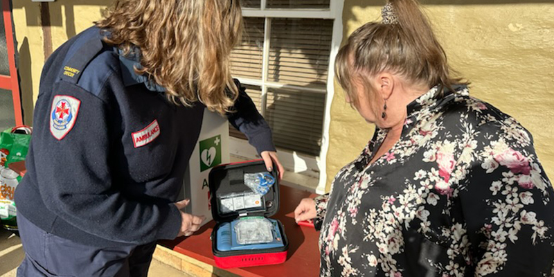 A paramedic is demonstrating how to use the automated external defibrillator device while a member of the Heathcote Community looks on.