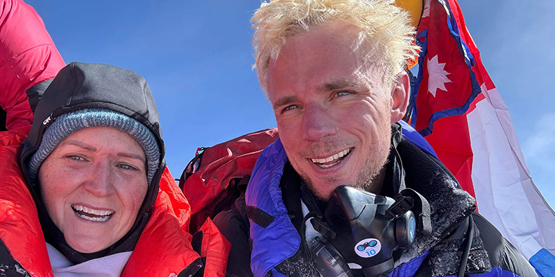 Two mountain climbers in winter gear smiling for the camera on a snowy mountain.