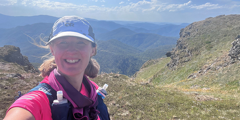 A female runner standing on an elevated rocky and hilly terrain with mountains in the background.