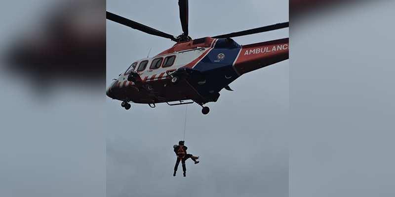 An aerial rescue operation with two people suspended from a helicopter hovering above ground.