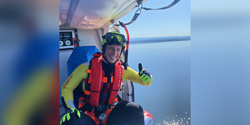 A man in a life jacket seated inside a helicopter flying over a lake.