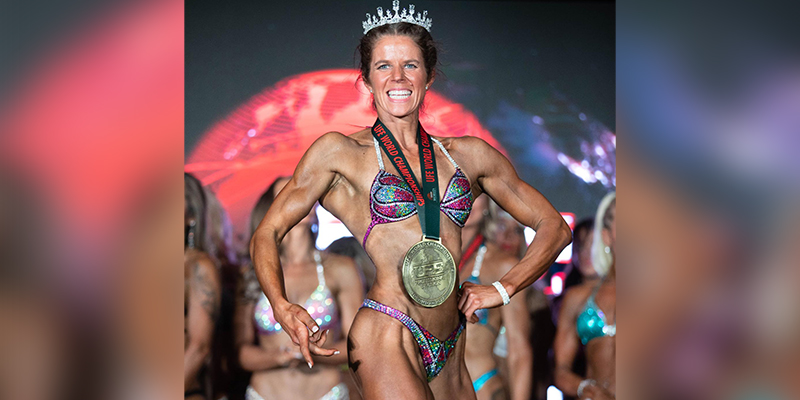 Female athlete in bikini proudly flexing her body muscles on stage with a medal around her neck and a tiara on her head.