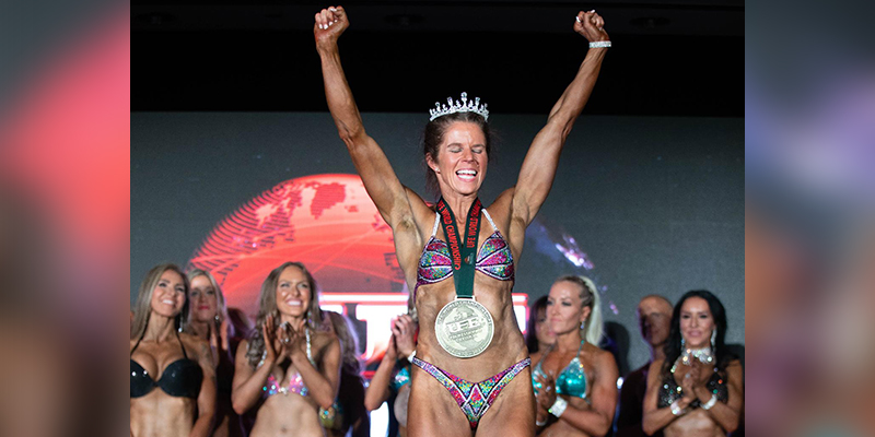 Female athlete in bikini with a medal around her neck and a tiara on her head raising her hands into the air.