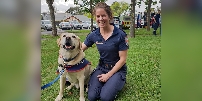 A female paramedic kneeling on the grass and petting a service dog.