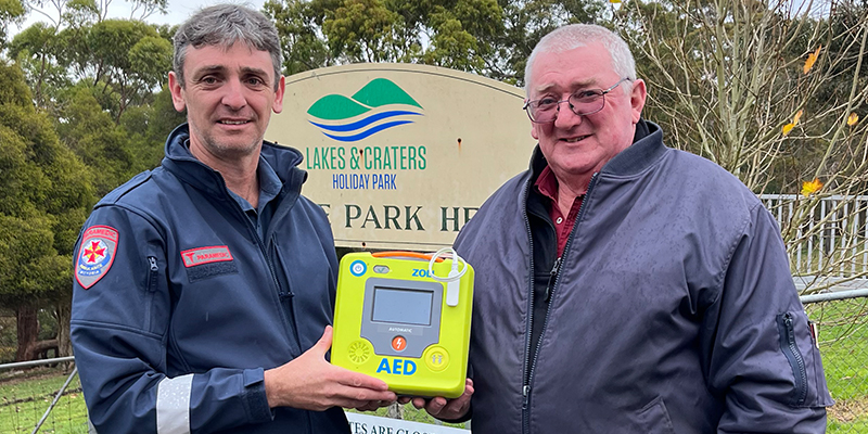 Two men standing in front of a sign while holding the Automated External Defibrillator device.