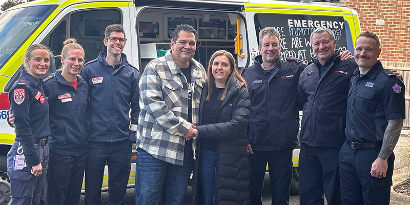 A man and a woman are holding hands, flanked by a group of paramedics by the side of an ambulance.