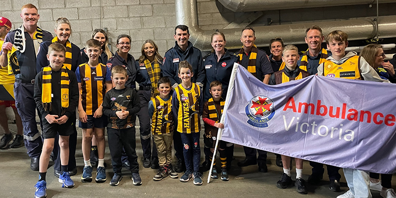 A diverse group of individuals comprising of adults and children stands together in front of a large Ambulance Victoria banner.