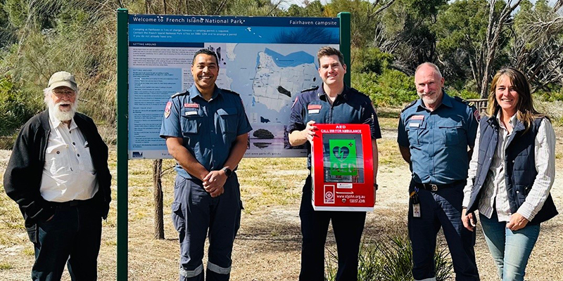 Cinq personnes se tiennent devant un panneau avec une carte dans une zone de camping. Une personne tient un défibrillateur externe automatisé.