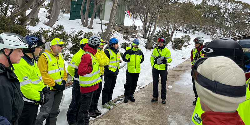 A group of men in snow and rescue gear gather around to discuss a plan on a snowy hillside.