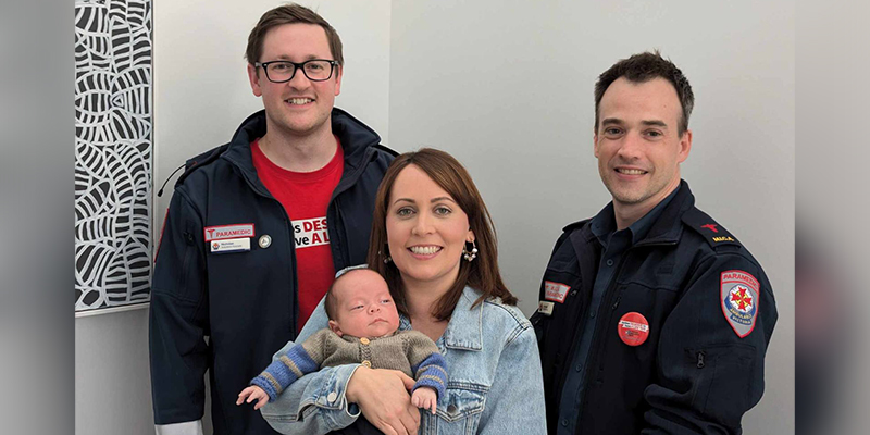 Two paramedics and a woman are standing in a room. The woman is holding an infant in her arms.