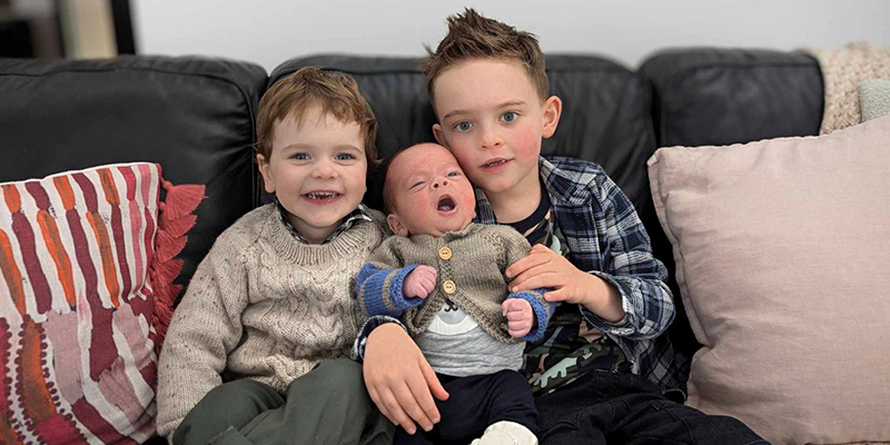 Two boys are sitting on a black sofa. One of the boys is holding an infant around his arms.