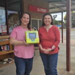 Two smiling women standing in front of a general store holding an AED.