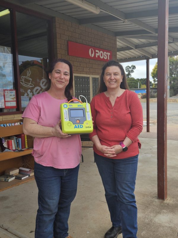 Two smiling women standing in front of a general store holding an AED.