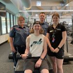 A teenage boy sits on a gym box holding a weight. Two paramedics and a gym staff member stand behind him, smiling for the camera.