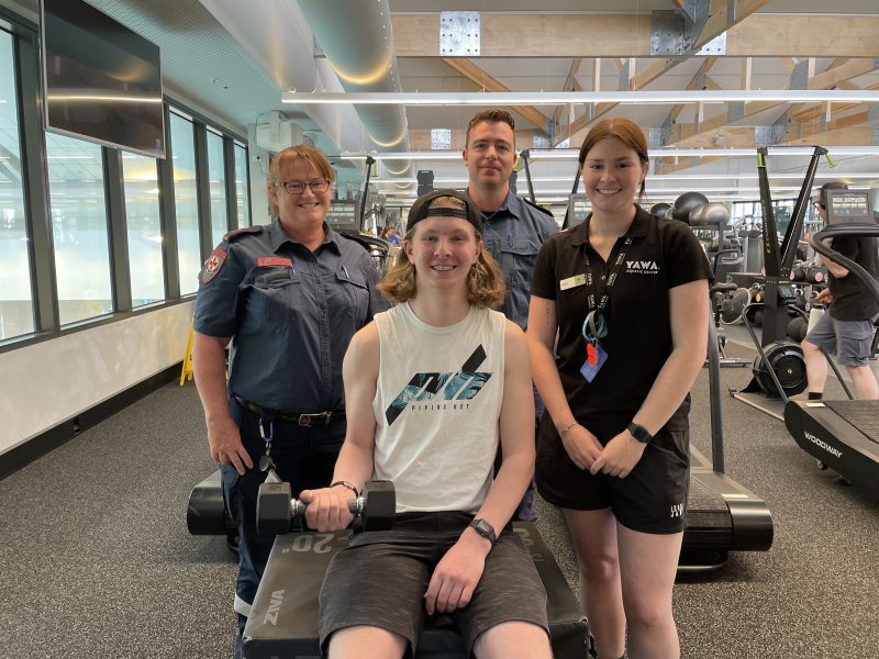 A teenage boy sits on a gym box holding a weight. Two paramedics and a gym staff member stand behind him, smiling for the camera.