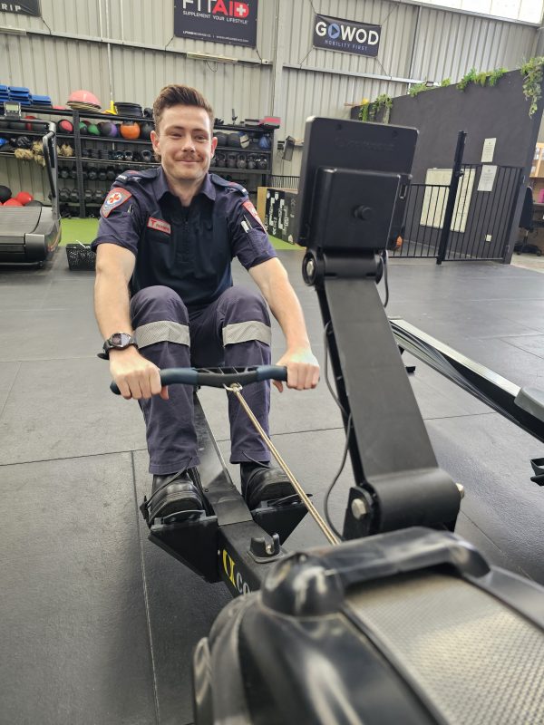 A man in Ambulance Victoria paramedic uniform on a rowing machine.