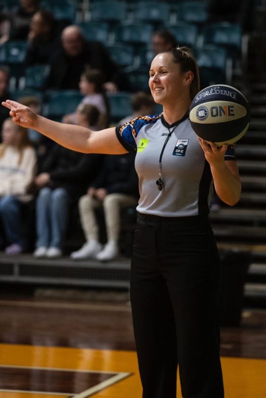 A woman referee holds the basketball and signals a call.