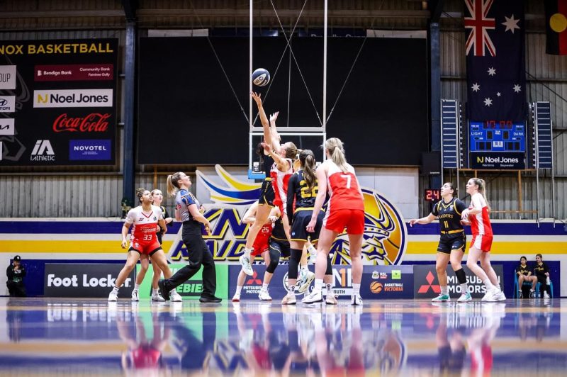 Woman basketball players jump for the ball as a woman referee moves out of the way.