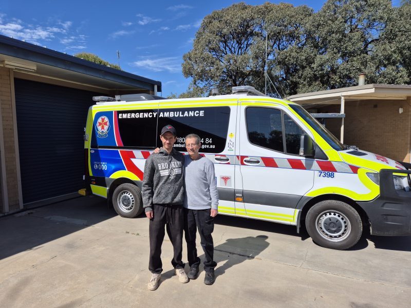 Two men with their arms around each other pose for the camera in front of an ambulance.