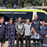 Ambulance Victoria paramedics and ACOs smile for the camera on either side of two men. The group of six stand in front of an ambulance.