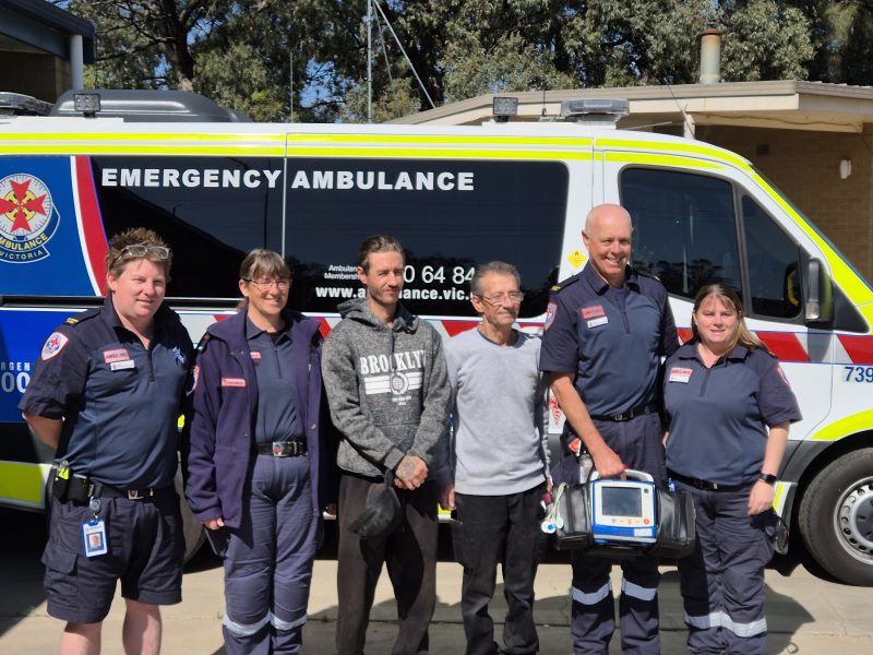 Ambulance Victoria paramedics and ACOs smile for the camera on either side of two men. The group of six stand in front of an ambulance.