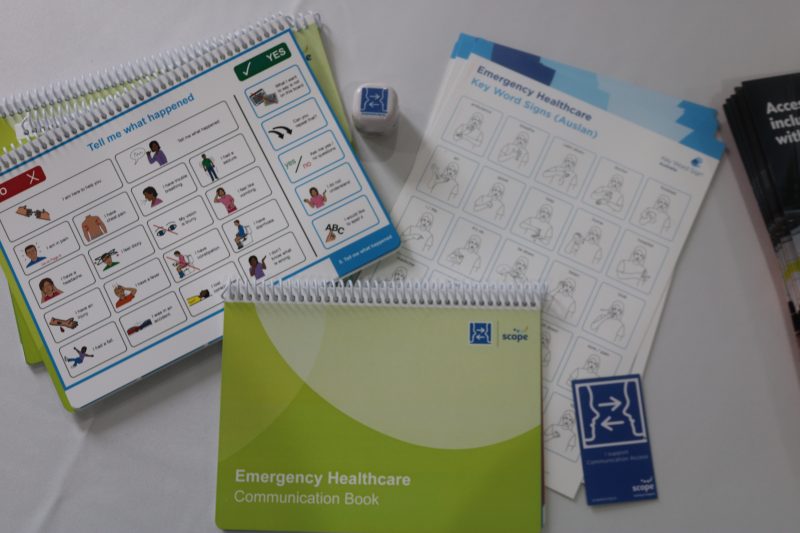 Books and information sheets are spread across a table. A book is titled 'Emergency Healthcare Communication Book'. There is an open copy which shows images and text to help someone describe what's wrong. There is also an information sheet titled 'Emergency Healthcare Key Word Signs (Auslan)' which shows diagrams of Auslan signs.