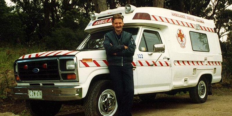 A man is leaning against the side of an old ambulance parked beside a row of trees.