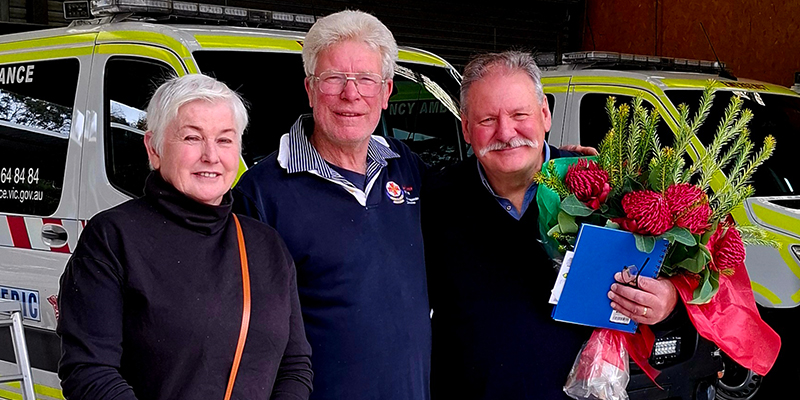 A woman and two men, one holding a bouquet of flowers, standing in front of two parked ambulances.