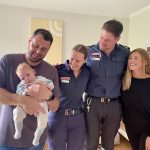 Four people stand in a line with their arms around each other. A smiling man holds a baby. Two paramedics look across and smile. A woman looks across and smiles as well.