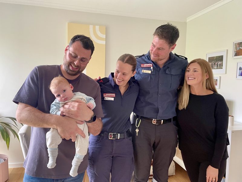 Four people stand in a line with their arms around each other. A smiling man holds a baby. Two paramedics look across and smile. A woman looks across and smiles as well.