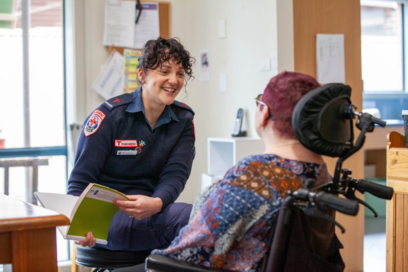 An Ambulance Victoria paramedic smiles towards a person in a wheelchair, who has their back to the camera.