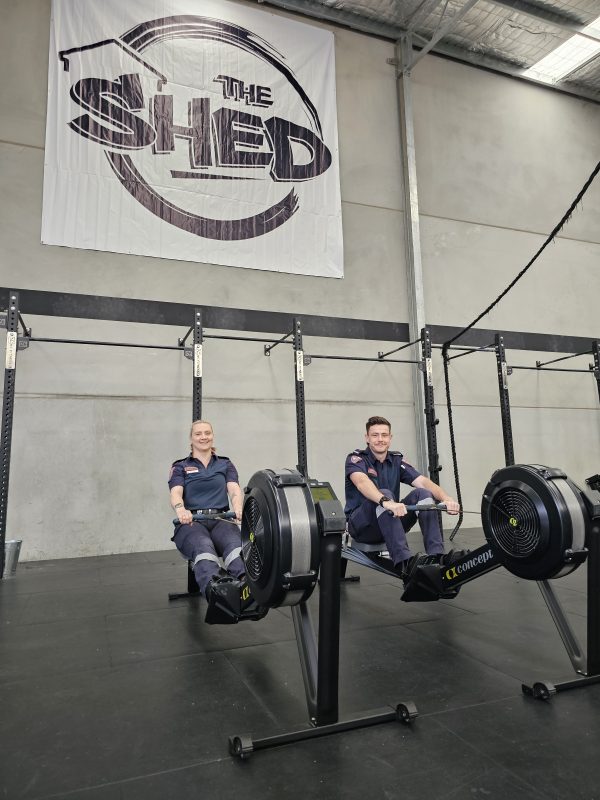 A man and woman in Ambulance Victoria paramedic uniform sit on rowing machines in The Shed Gym.