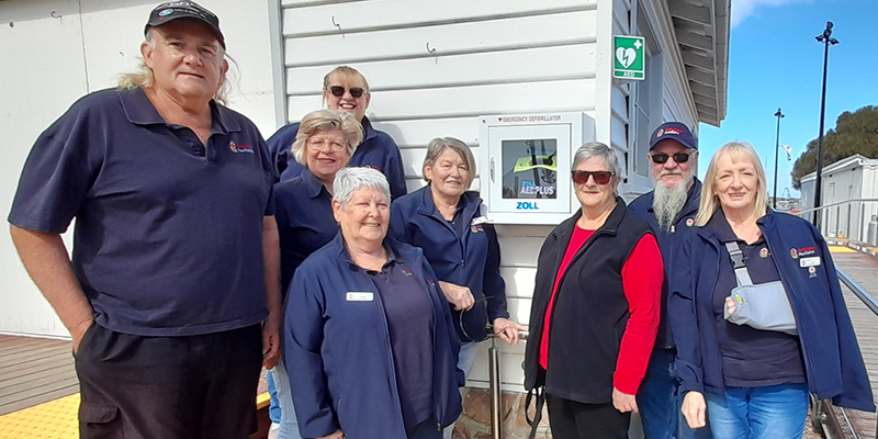 A group of people standing beside a building. There is a automated external defibrillator device attached to the wall of the building.