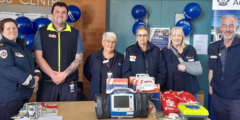 A group of people standing behind a table. There is a automated external defibrillator device on the table.