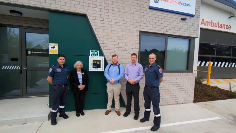 A group of five people stand next to an AED mounted on the wall of an Ambulance Victoria branch. Two of the group are paramedics.