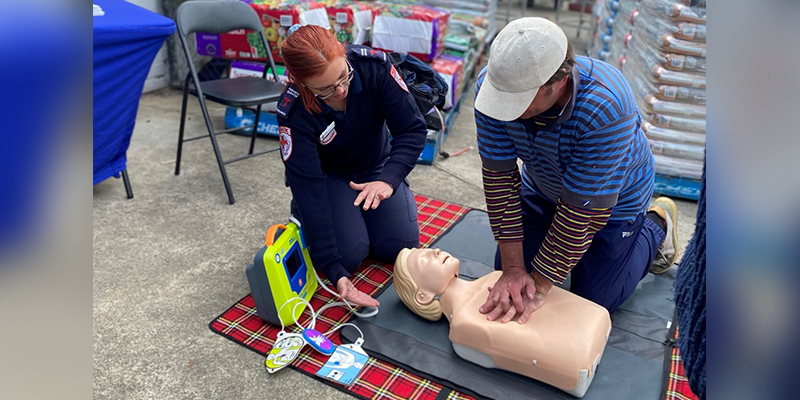 A man is performing cardiopulmonary resuscitation on a manikin. A female paramedic is providing guidance beside the man.