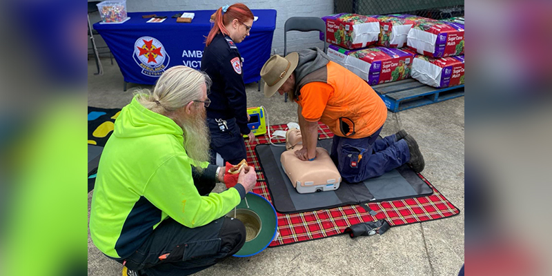 A man is performing cardiopulmonary resuscitation on a manikin while a female paramedic and another man are observing from the side. 