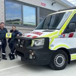 Two paramedics stand next to an ambulance holding an AED.