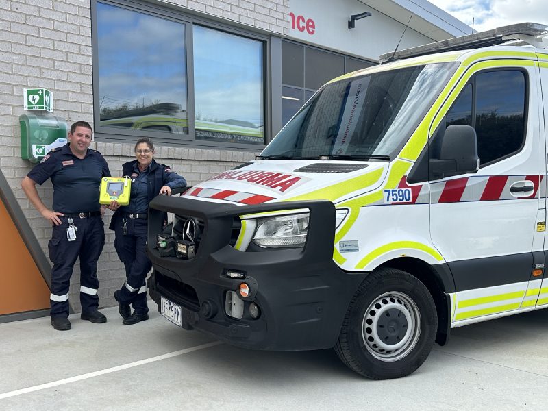 Two paramedics stand next to an ambulance holding an AED.