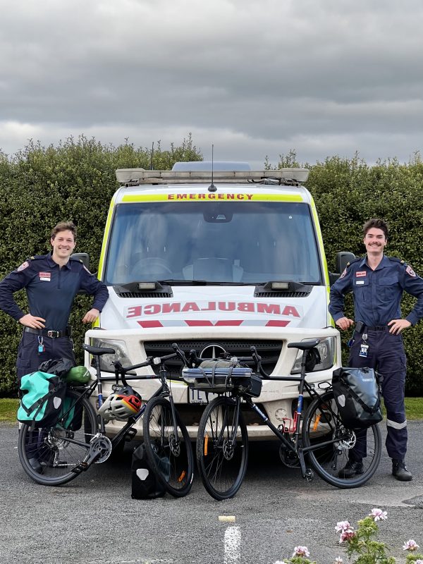 Two men in Ambulance Victoria on-road uniform smile for the camera, standing either side of an ambulance. Their bicycles are parked in front of the ambulance.