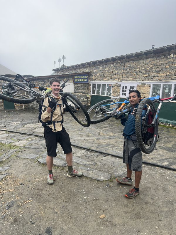 Two men hold mountain bikes over their shoulders and smile for the camera in front of Throng High Camp Restaurant. It is cloudy.