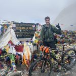 A man with a mountain bike smiles for the camera in front of a sign marking the highest point on the Annapurna Circuit - the Thorang-La Pass, 5416m. Colourful prayer flags surround the sign. It is cloudy.