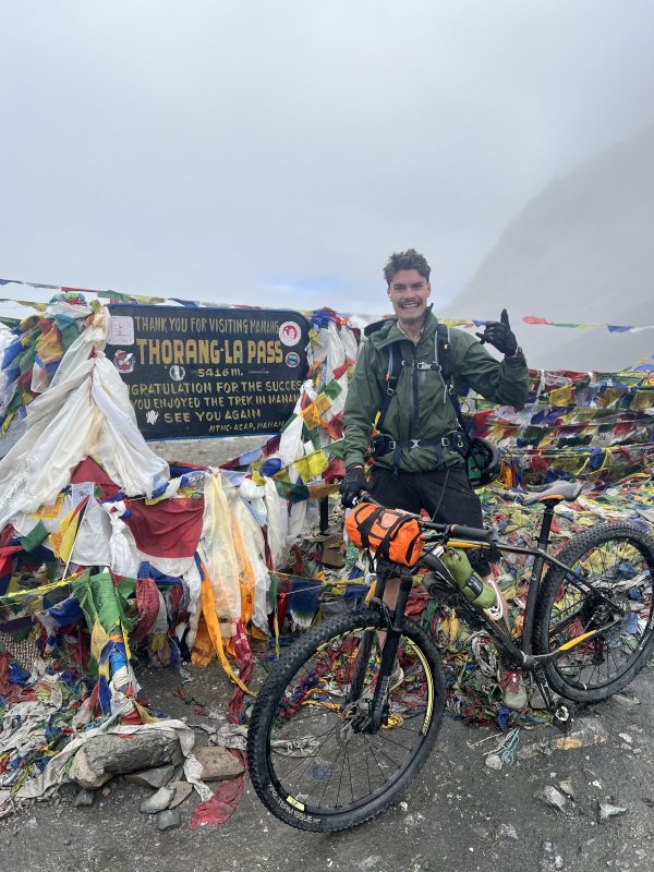 A man with a mountain bike smiles for the camera in front of a sign marking the highest point on the Annapurna Circuit - the Thorang-La Pass, 5416m. Colourful prayer flags surround the sign. It is cloudy.