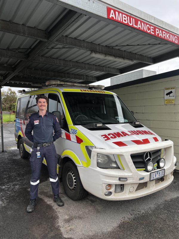 A man in Ambulance Victoria on-road uniform smiles for the camera, standing in front of a parked ambulance.